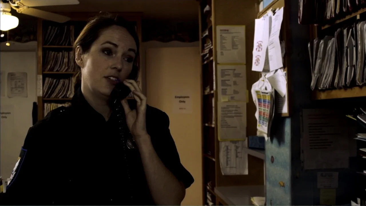 Abandoned Dead (2015) movie still: medium — A cramped office workspace is cluttered with stacked files on metal shelving units. A woman in a dark…