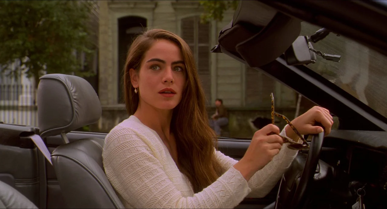 Hard Target (1993) movie still: medium — A woman sits in the driver seat of a convertible with grey leather upholstery. She holds a pair of tortoise…