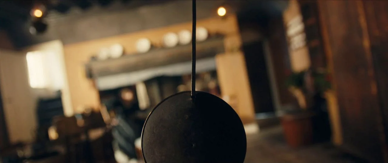 The Personal History of David Copperfield (2019) movie still: closeup — A dark metal pendulum hangs in the foreground of a blurred domestic space.…