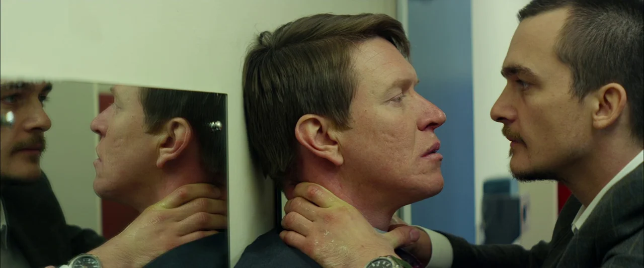 Starred Up (2013) movie still: medium — A tiled public restroom with a visible mirror surface reflecting the scene. One man presses another man…
