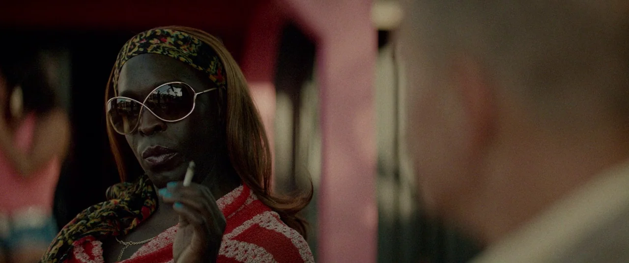 Triple 9 (2016) movie still: medium — A woman stands in a bright urban area near a pink building structure. She wears large sunglasses and holds a…