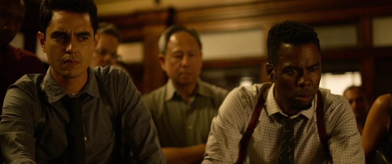 Spiral (2021) movie still: medium — A dimly lit room features wood-paneled walls and several men gathered in the workspace. Two men in the…
