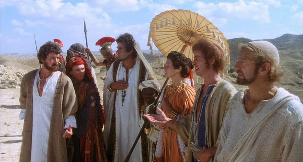 Life of Brian (1979) movie still: fullbody — A group of figures in ancient-style robes gather on a sandy, arid hillside. A large yellow parasol is…