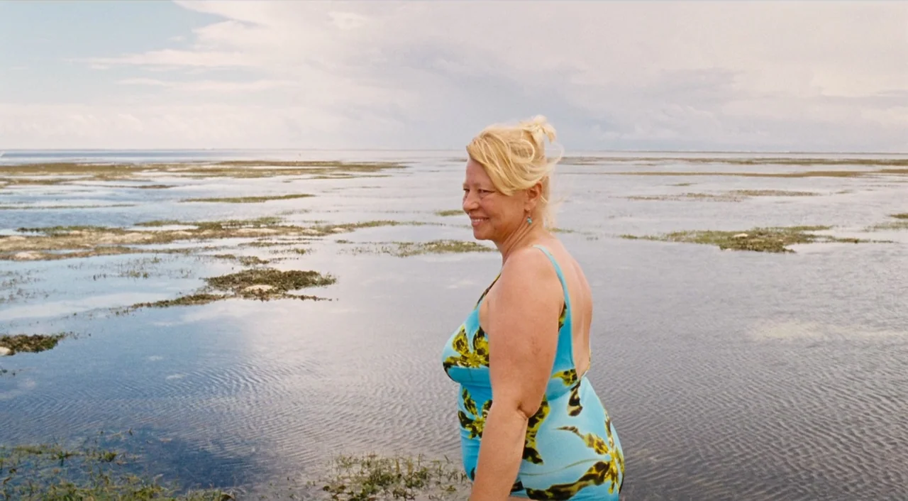Paradise: Love (2012) movie still: medium — A woman stands waist-deep in shallow coastal water under an overcast sky. The water is calm with patches…