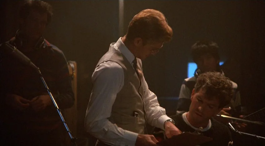 The Coca-Cola Kid (1985) movie still: medium — A dimly lit recording studio is filled with audio equipment and musical instruments. A man holding a…