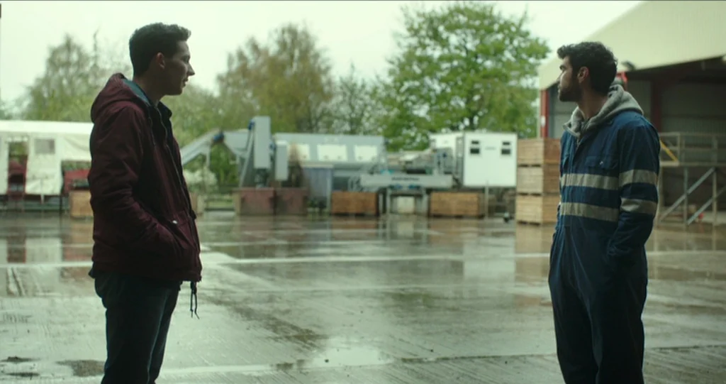 God’s Own Country (2017) movie still: fullbody — An open concrete yard of an industrial site features heavy machinery and wooden crates. Two young…