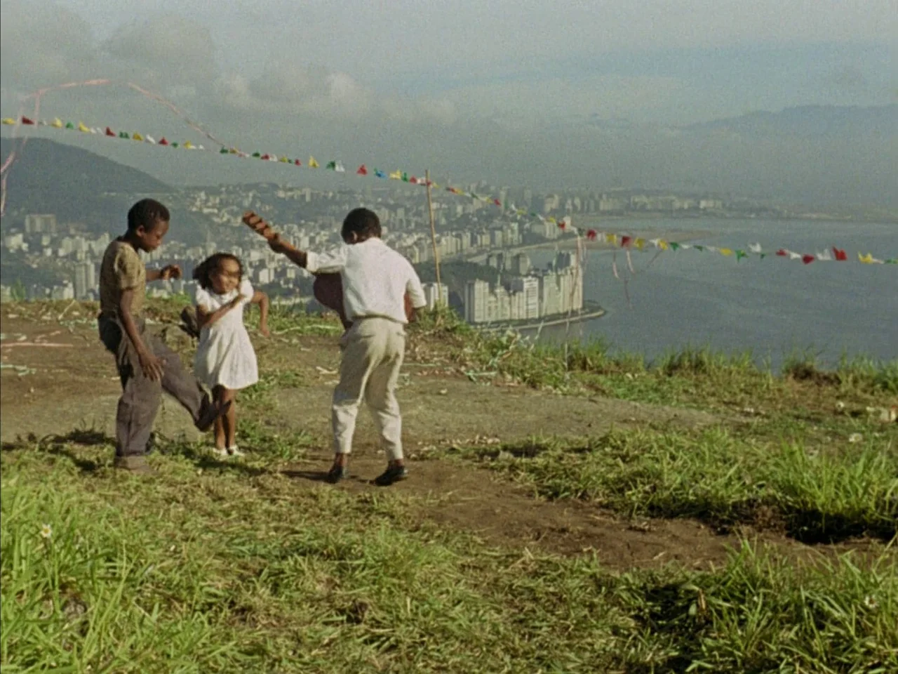 Black Orpheus (1959) movie still: fullbody — A grassy, unpaved hillside overlooking a coastal city skyline and large bay. Three children dance…
