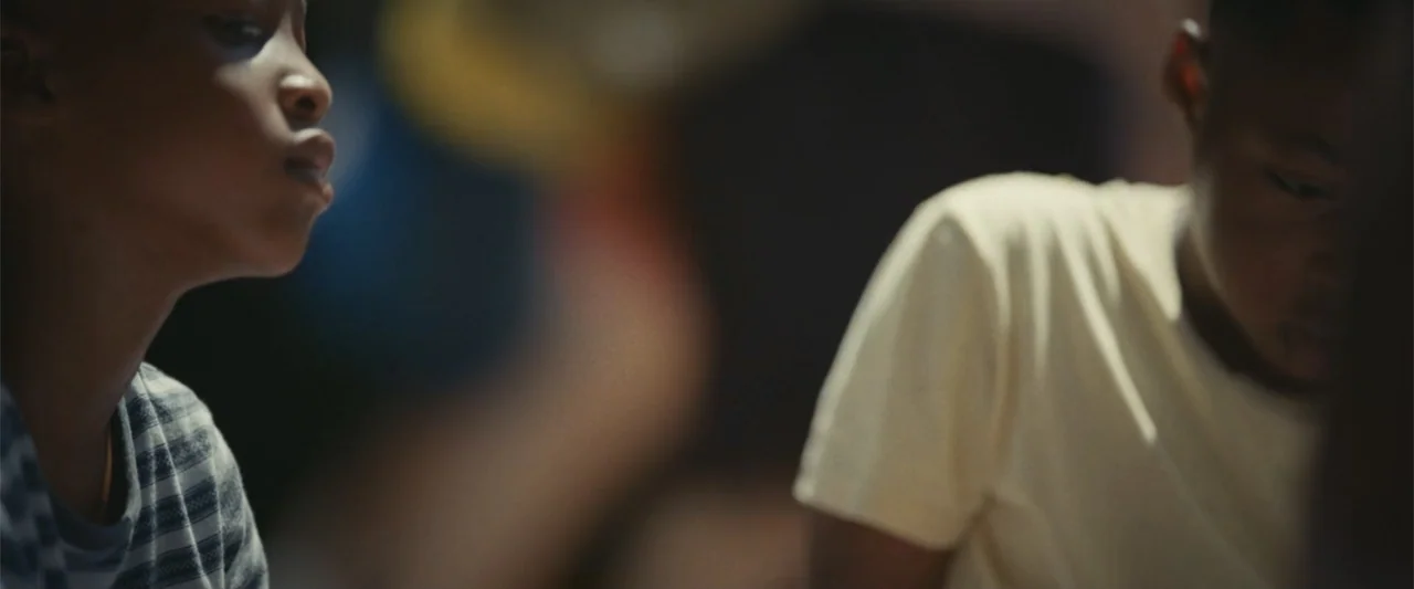 Giannis: The Marvelous Journey (2024) movie still: medium — A dimly lit indoor space with two boys positioned in the foreground. One boy wears a…