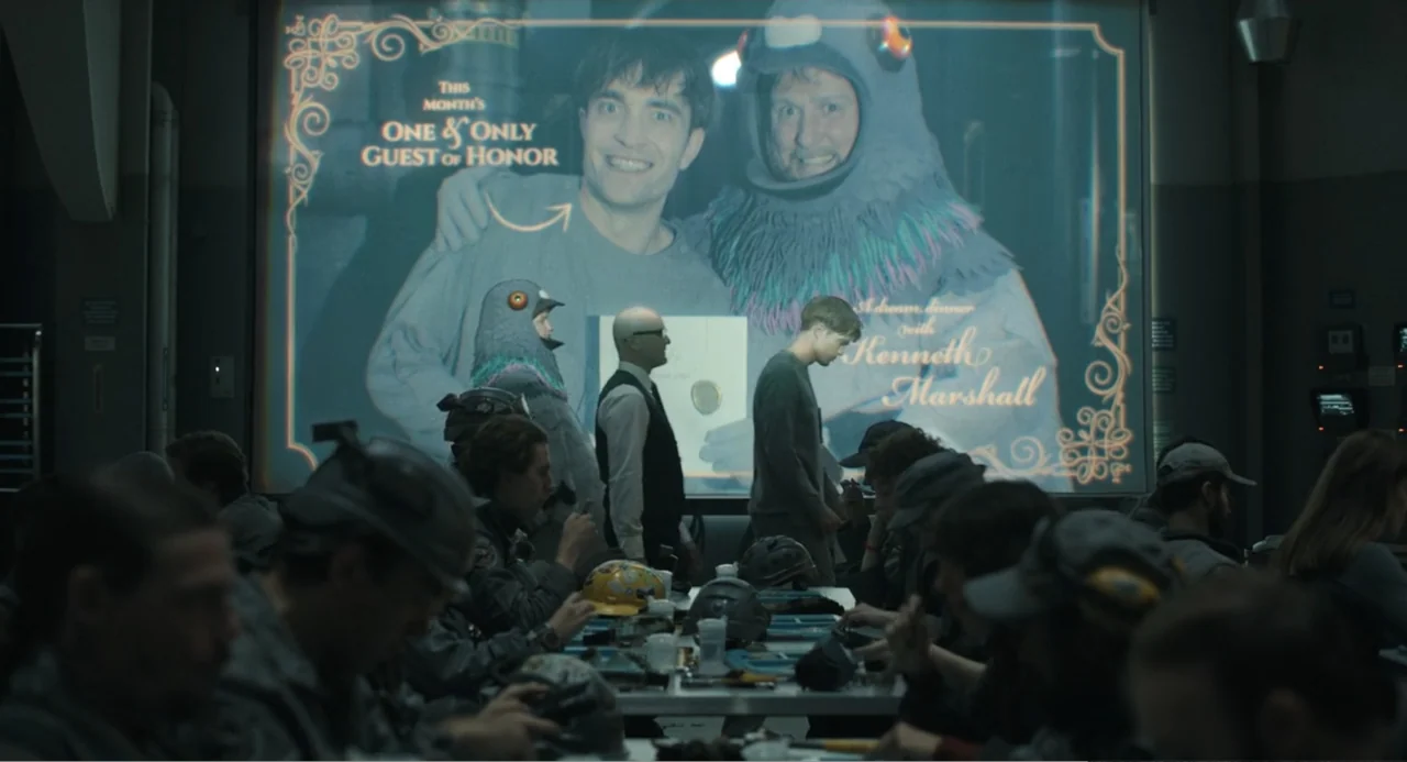 Mickey 17 (2025) movie still: wide — A dimly lit communal room contains long tables filled with people in grey uniforms. A large projection on the…