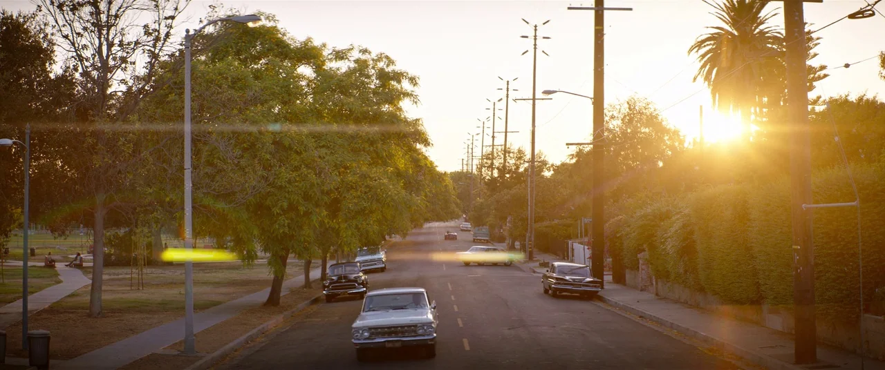 Ford V Ferrari (2019) movie still: wide — A quiet suburban road is lined with mature trees and vintage cars at sunset. The setting sun causes…