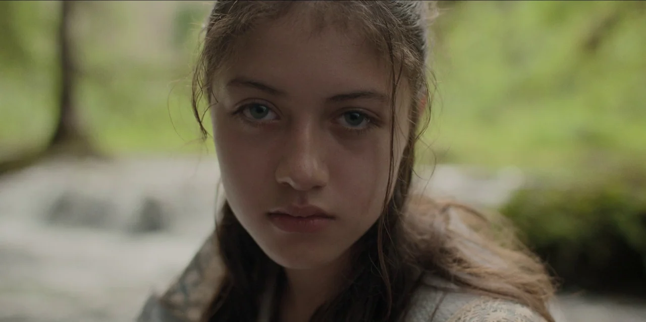 Pedro Páramo (2024) movie still: closeup — A natural, outdoor setting features a flowing water source behind the subject. A teenage girl looks…