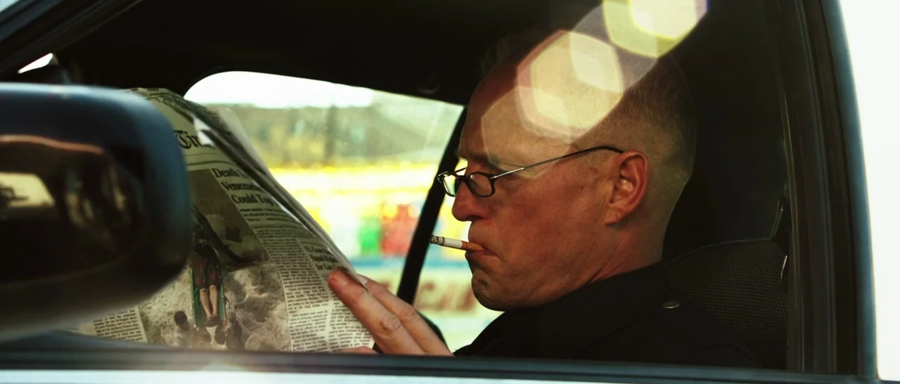 Rampart (2011) movie still: closeup — A man wearing a uniform sits inside a vehicle while reading a newspaper. Sunlight reflects off the car window…