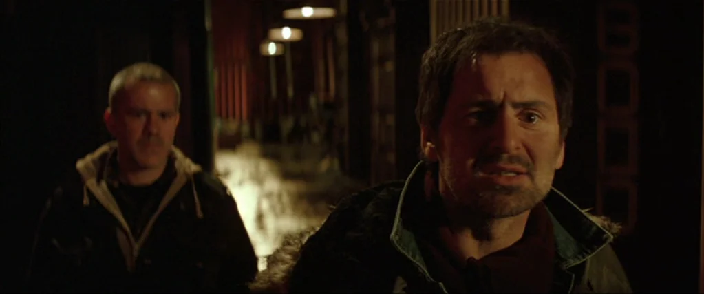Batman Begins (2005) movie still: medium — Two men stand in a darkened, narrow corridor with warm lights visible in the distance. The foreground man…