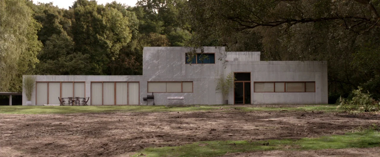 Borgman (2013) movie still: wide — A modern, boxy concrete house with floor-to-ceiling windows sits on a large, patchy lawn. Dense green trees…