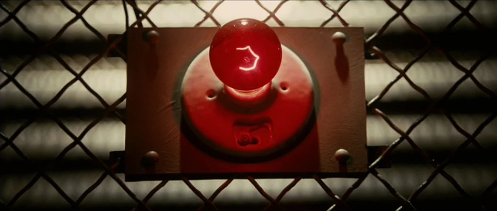 Shutter Island (2010) movie still: closeup — A bright red glowing light bulb is mounted against a dark industrial surface. Chain-link wire fencing…