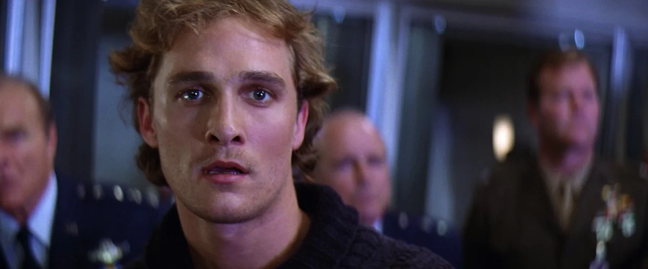 Contact (1997) movie still: closeup — A dimly lit room with several individuals in military uniforms visible in the background. A young man in a…