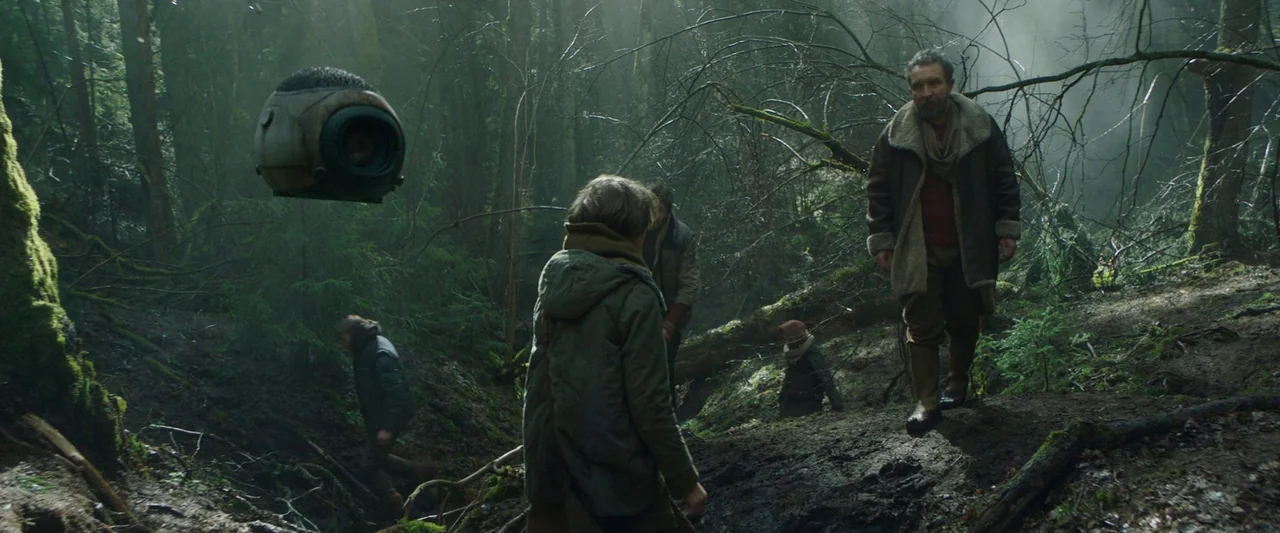 Vesper (2022) movie still: wide — A dense, overgrown temperate forest with damp soil, fallen logs, and thick light fog. A group of people hike…