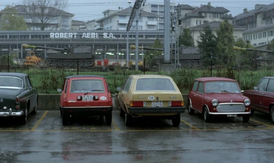 Every Man for Himself (1980) movie still: wide — A row of parked vintage cars sits in a wet, paved parking lot. Behind the parking area, large…