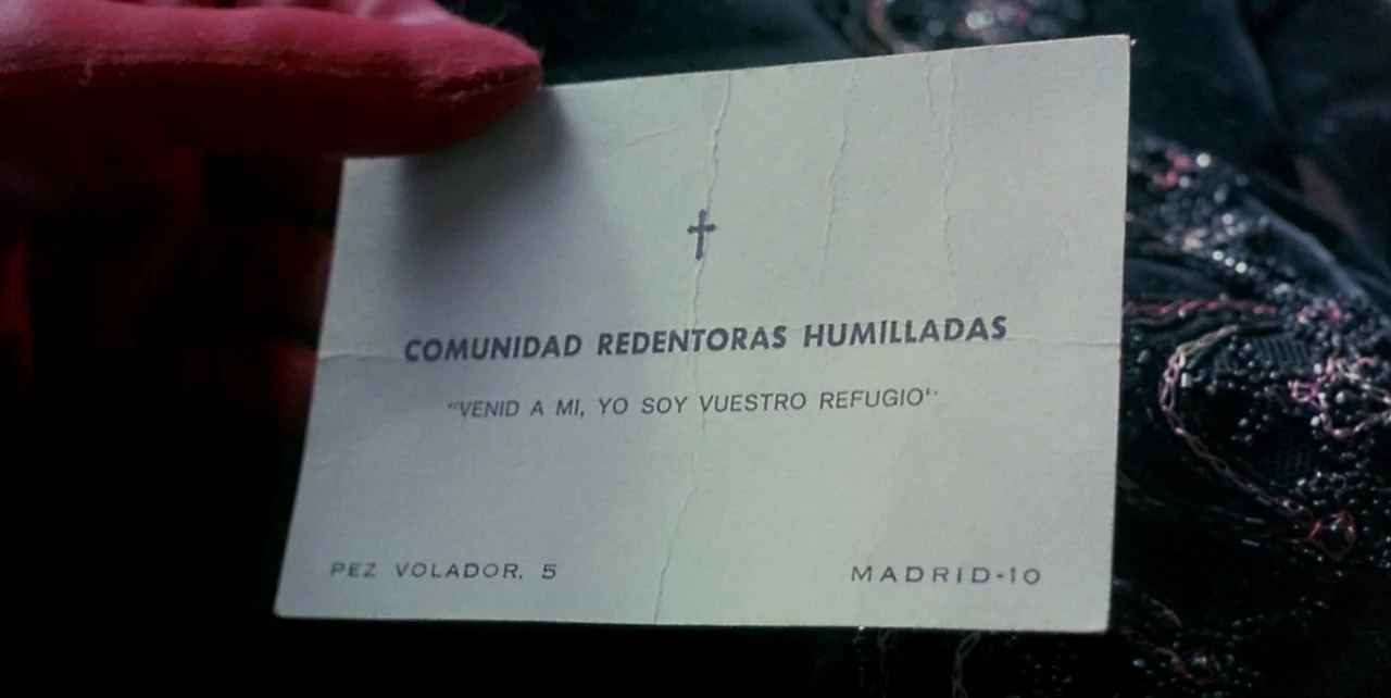 Dark Habits (1983) movie still: closeup — A close-up of a printed paper card featuring religious text and a Madrid address. The card is held by a…