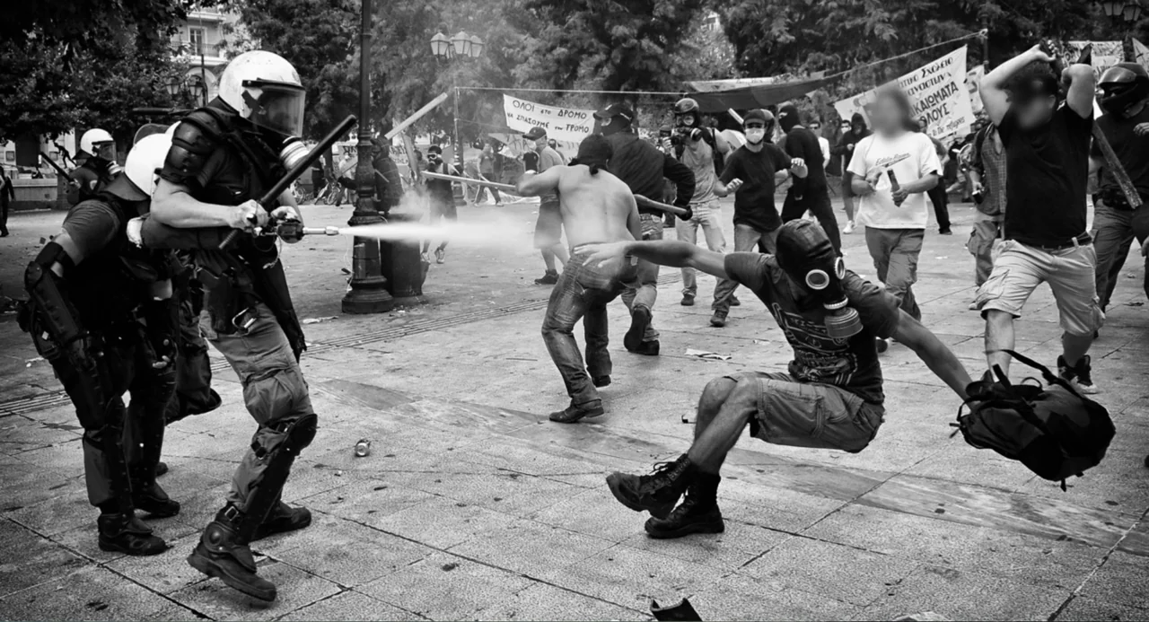 Az X- Kryptonit projekt (2020) movie still: fullbody — An open plaza filled with protestors and riot police during a civil disturbance. A police…