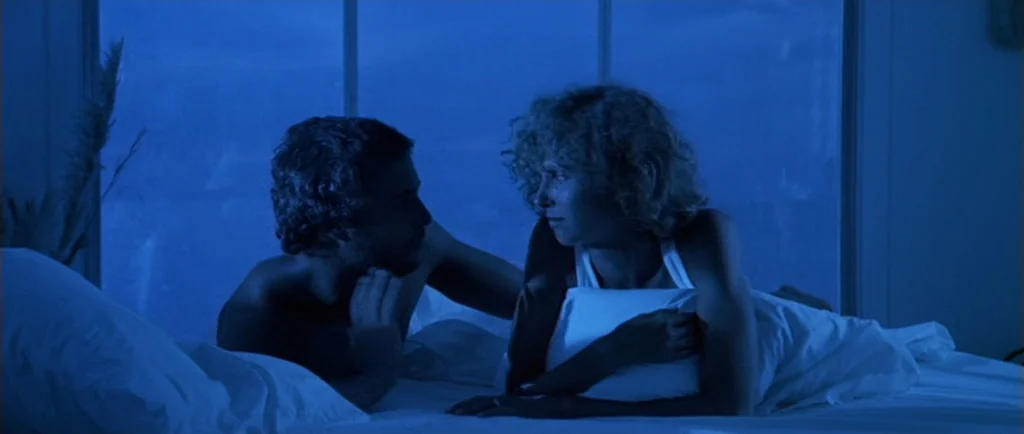 Manhunter (1986) movie still: medium — A man and woman lie in a bed positioned against a large glass window. The room is bathed in deep blue light…