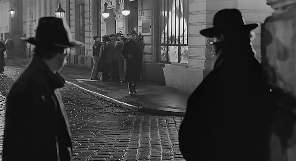 Kafka (1991) movie still: medium — A nighttime cobblestone street features two men in the foreground watching a distant group. The men stand in the…