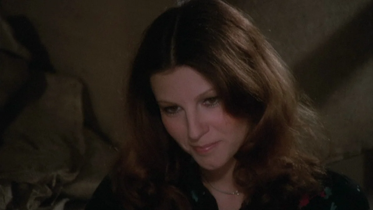 The Black Belly of the Tarantula (1971) movie still: closeup — A dimly lit interior space with indistinct dark textures in the background. A young…