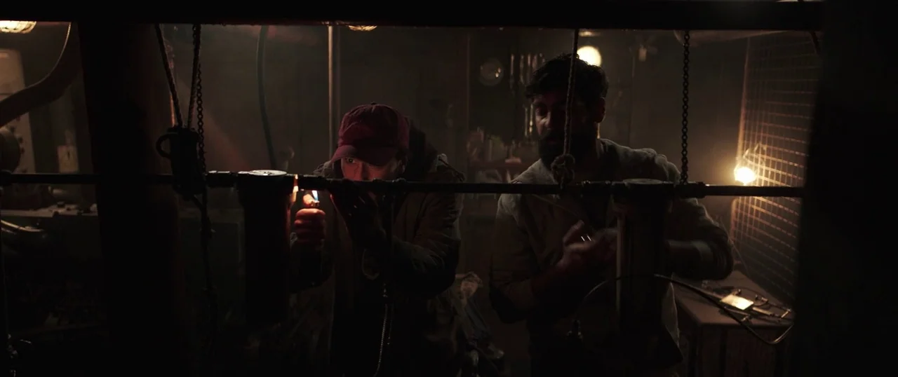 Sea Fever (2019) movie still: medium — A cluttered, dark, industrial subterranean space is filled with pipes and machinery. Two men perform repairs…