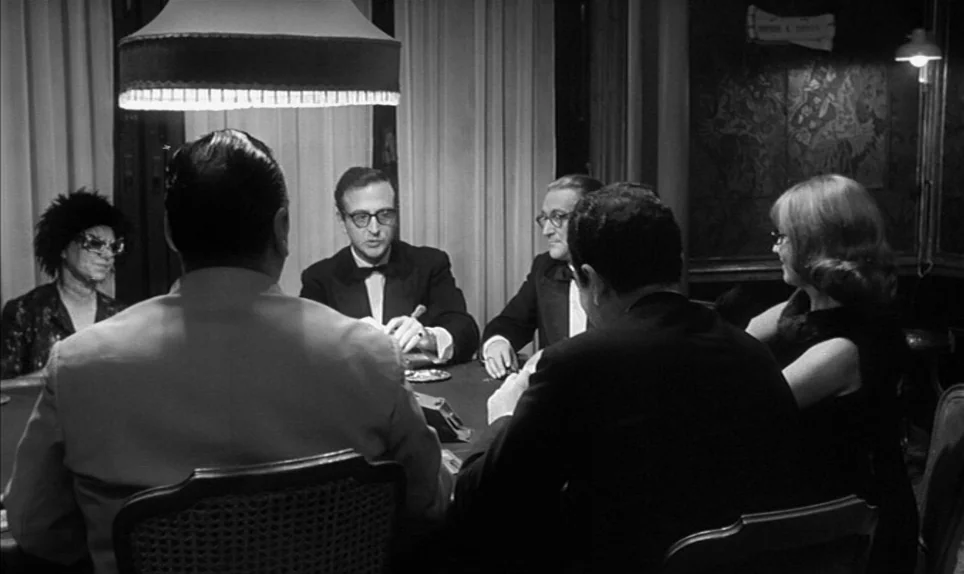 Eva (1962) movie still: medium — A group of six people sit around a card table in a dimly lit room. They are engaged in a game of cards under a…
