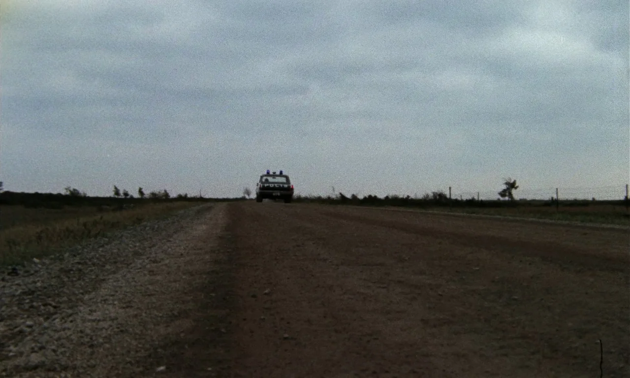 Thriller: A Cruel Picture (1973) movie still: wide — A dark police cruiser travels away down a long, desolate, unpaved rural road. The flat horizon…