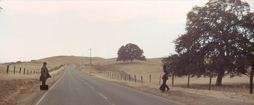 Scarecrow (1973) movie still: wide — An asphalt road cuts through dry, rolling hills under an overcast sky. Two men stand on opposite shoulders of…