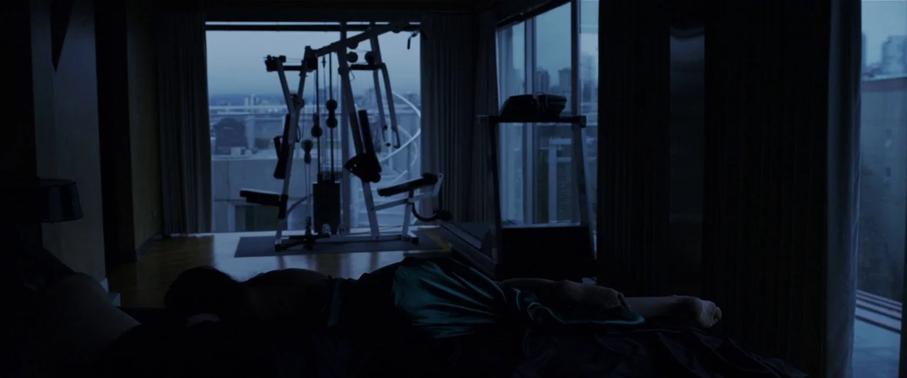 American Mary (2012) movie still: medium — A modern room features a dark sleeping area connected to an exercise space with equipment. A person lies…
