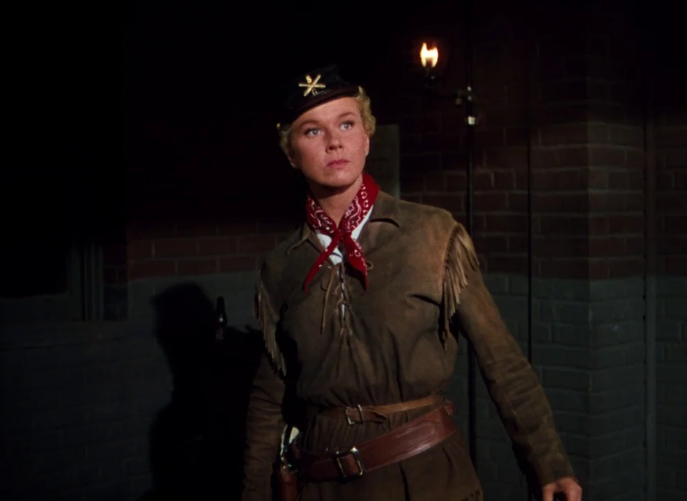 Calamity Jane (1953) movie still: medium — A woman stands before a dark brick wall at night lit by a single lantern. She wears a leather jacket and…