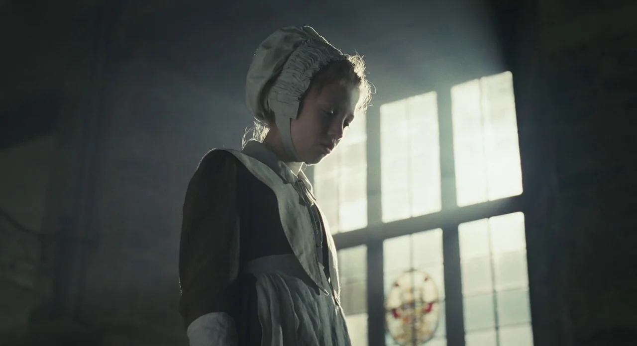 Jane Eyre (2011) movie still: medium — The interior of a church with stone walls and a tall, multi-pane glass window. A young girl in a period…