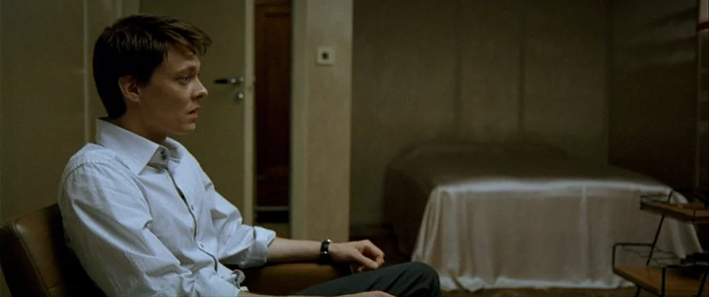Next Door (2005) movie still: medium — A dimly lit room features a neatly made bed and a leather chair. A young man sits in the chair looking…