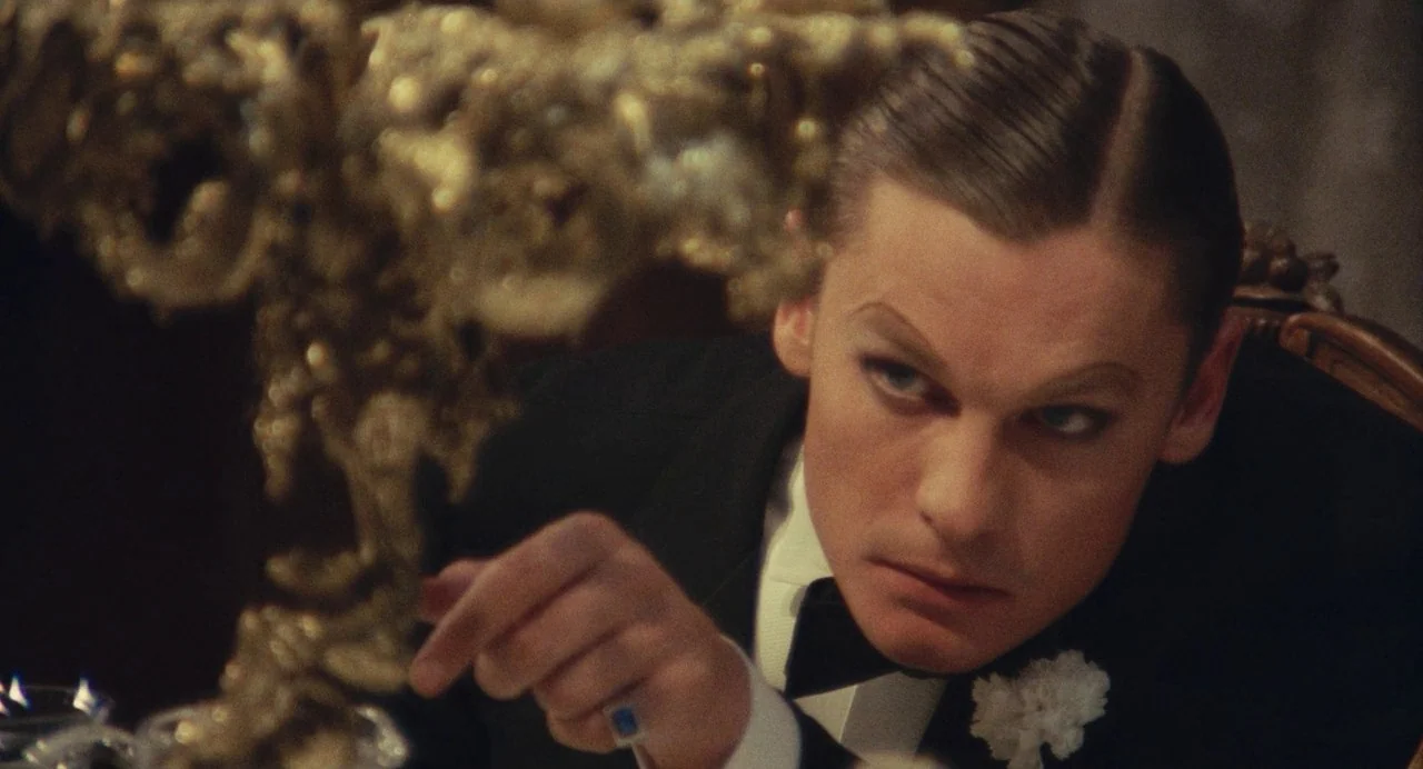 The Damned (1969) movie still: closeup — A dimly lit, formal interior space featuring an ornate gold candelabra in the foreground. A man in a tuxedo…
