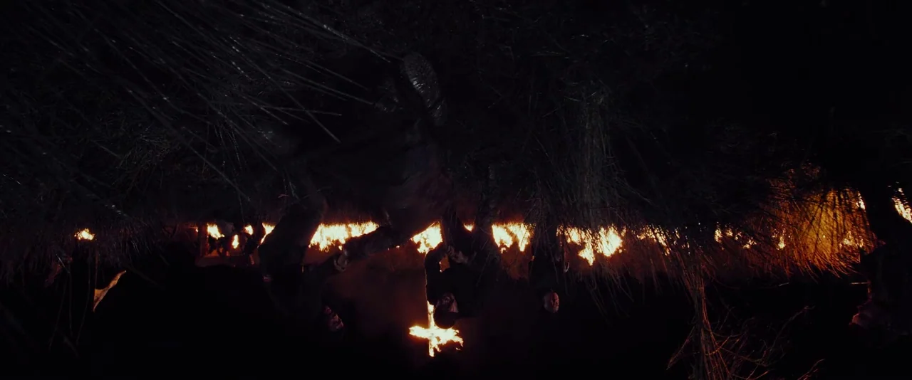 Apostle (2018) movie still: wide — A field at night features a large burning cross and a line of fire. Several figures stand in the dark grass,…