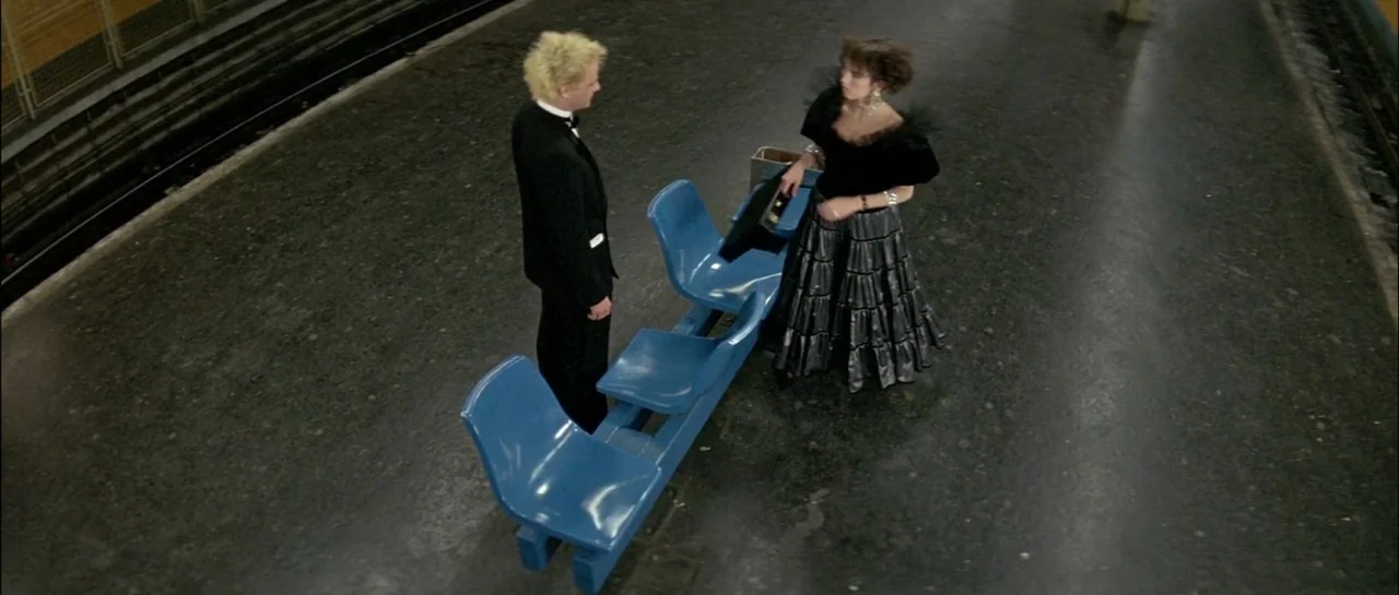 Subway (1985) movie still: wide — A desolate subway platform features a dark grey floor and blue molded plastic seating. A man and a woman in…