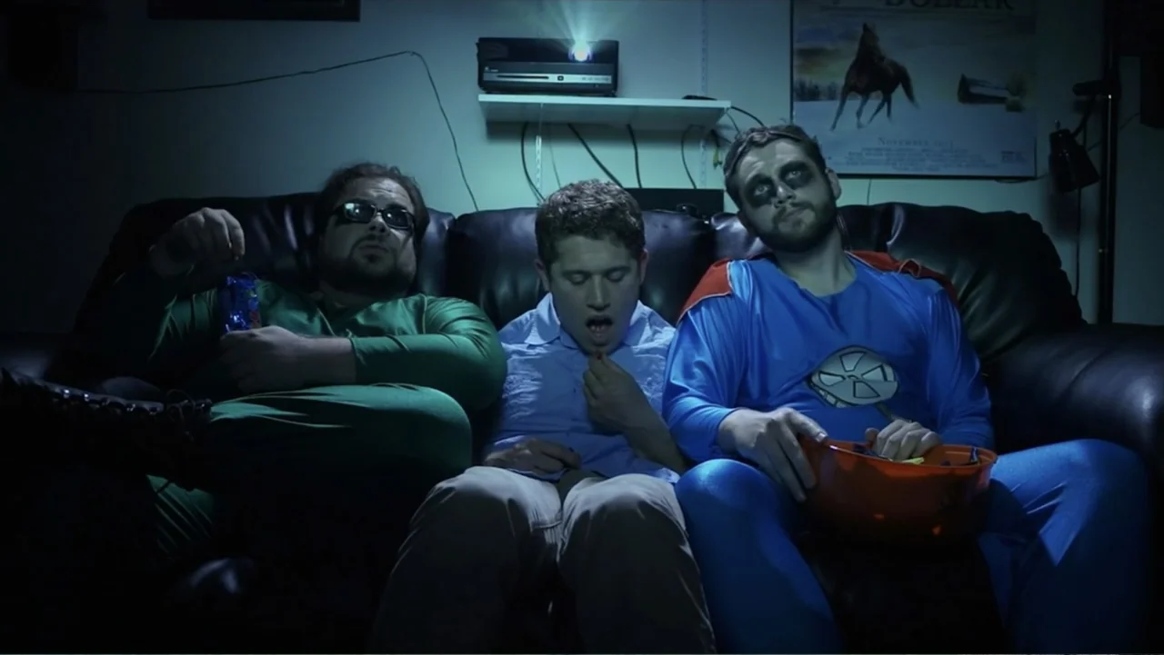 Hello I Love You (2018) movie still: medium — A dark, dimly lit room contains a black leather couch occupied by three men. The men sit together…