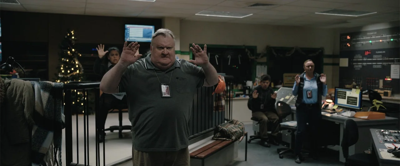 Play Dirty (2025) movie still: medium — A dimly lit industrial control room contains technical consoles and a lit Christmas tree. An older man…