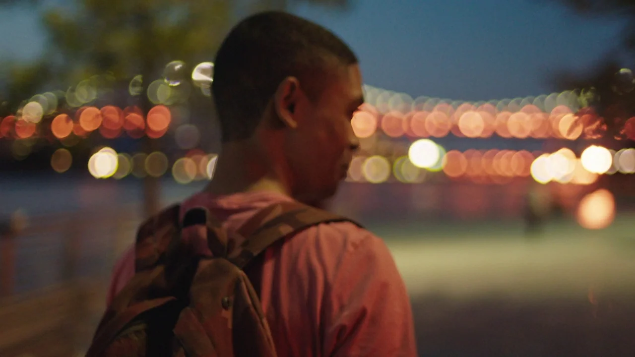 Madeline’s Madeline (2018) movie still: medium — A nighttime waterfront promenade features a blurred expanse of city bridge lights behind. A young…