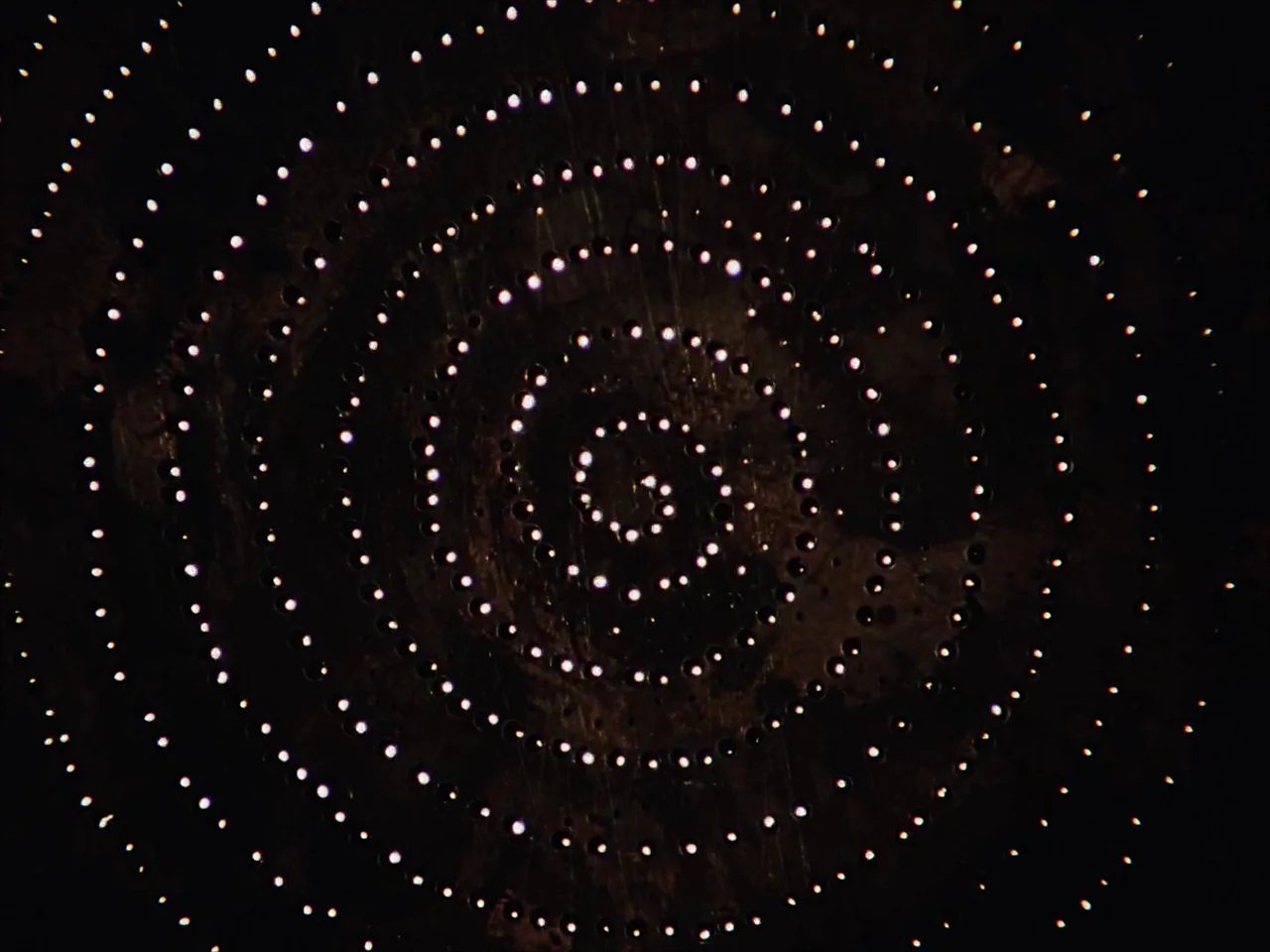 Kin-dza-dza! (1986) movie still: closeup — A dark surface is perforated with numerous small holes arranged in a tight spiral. Bright white light…