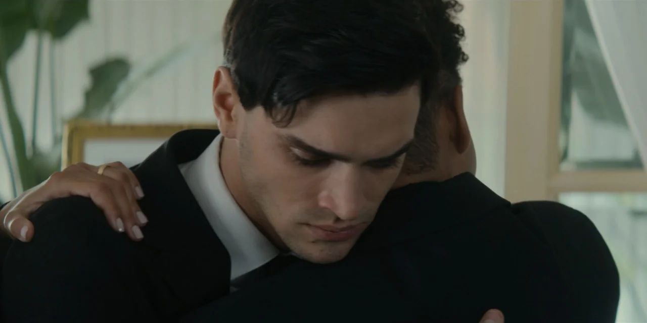 Our Fault (2025) movie still: closeup — An indoor living space with blurred white curtains visible in the background. A man in a suit jacket…