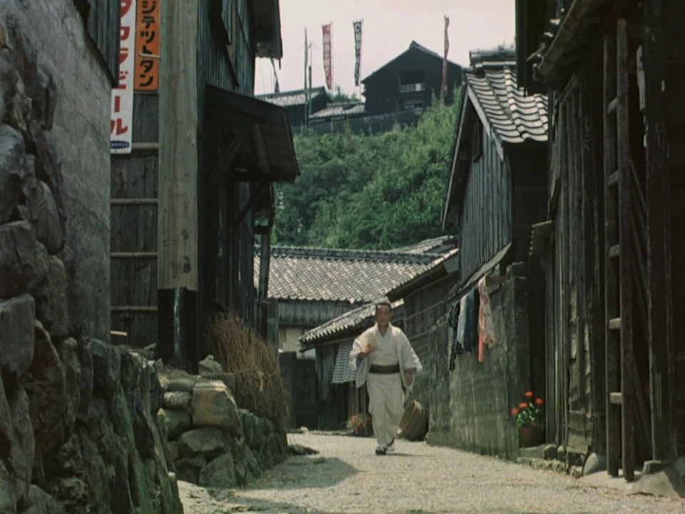Floating Weeds (1959) movie still: wide — A narrow Japanese village alleyway flanked by weathered wooden buildings and stone retaining walls. A man…