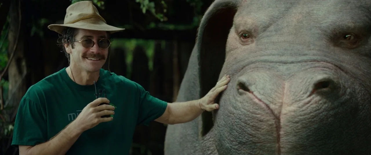 Okja (2017) movie still: medium — A dense green environment contains a man standing next to a large creature. The man wears a fedora and t-shirt…