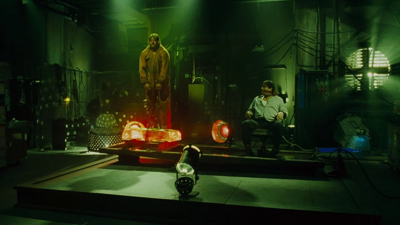 Saw IV movie still: wide — A dark industrial workshop contains electrical wiring, metal structures, and various mechanical tools. A man hangs in…