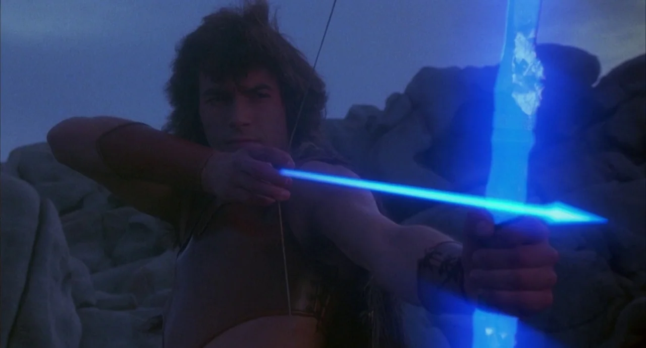 Conquest (1983) movie still: medium — A rocky, barren landscape provides the backdrop for a figure in fantasy armor. A young man aims a glowing neon…