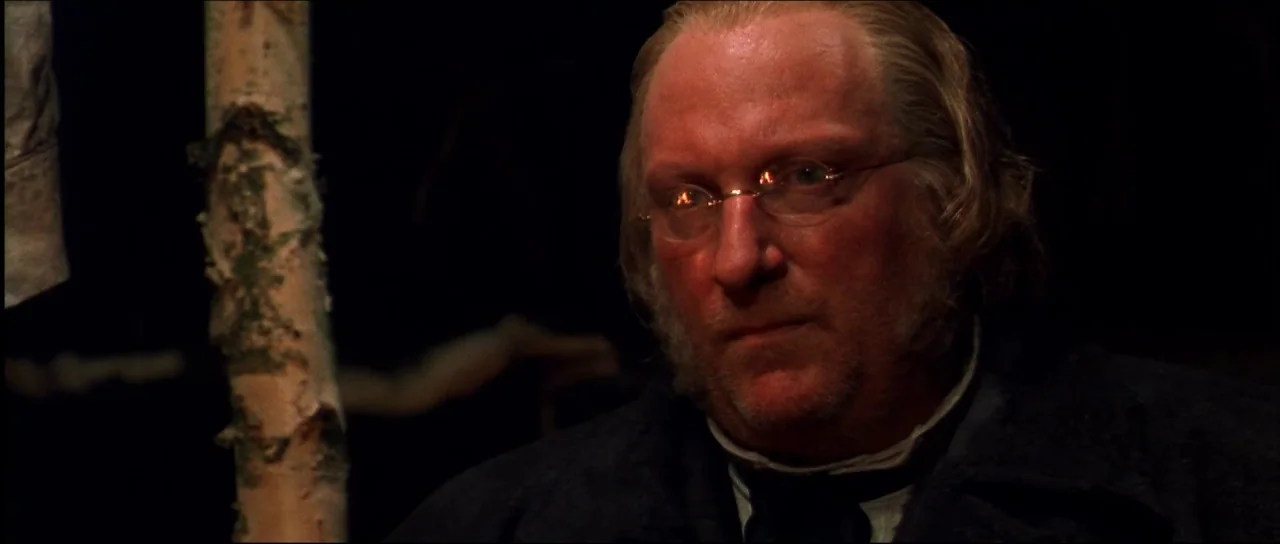 Ravenous (1999) movie still: closeup — A dark night setting featuring a birch tree trunk in the foreground. An older man wearing spectacles looks…
