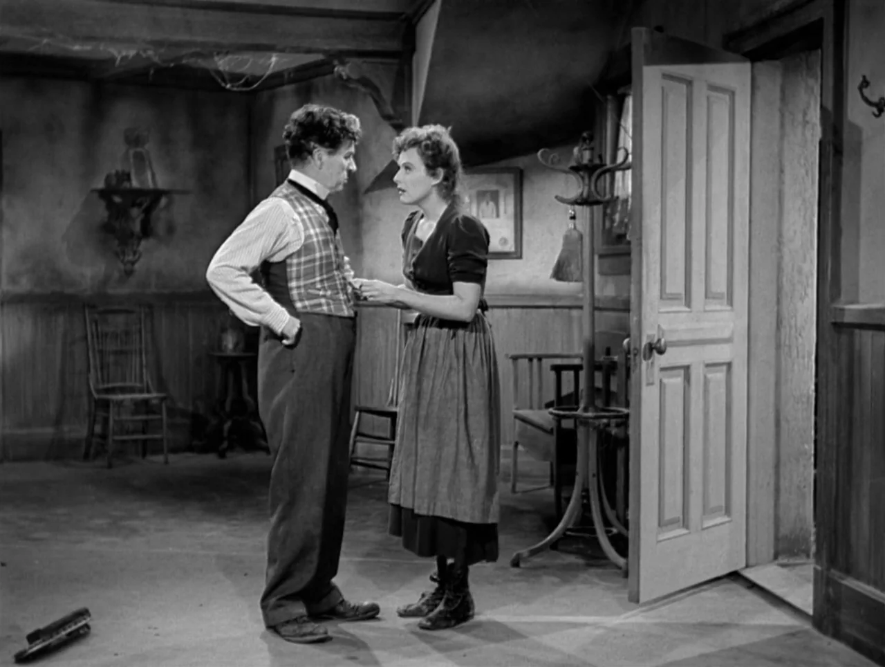 The Great Dictator (1940) movie still: fullbody — A rustic, sparsely furnished room features wood-paneled walls and an open doorway. A man and woman…