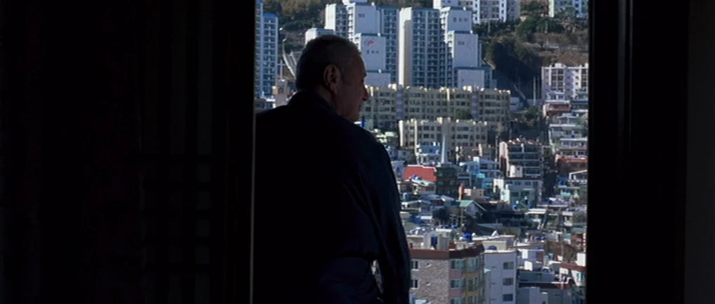 The Intruder (2004) movie still: medium — An elderly man is framed by a dark window looking out at a city. Densely packed high-rise apartment…